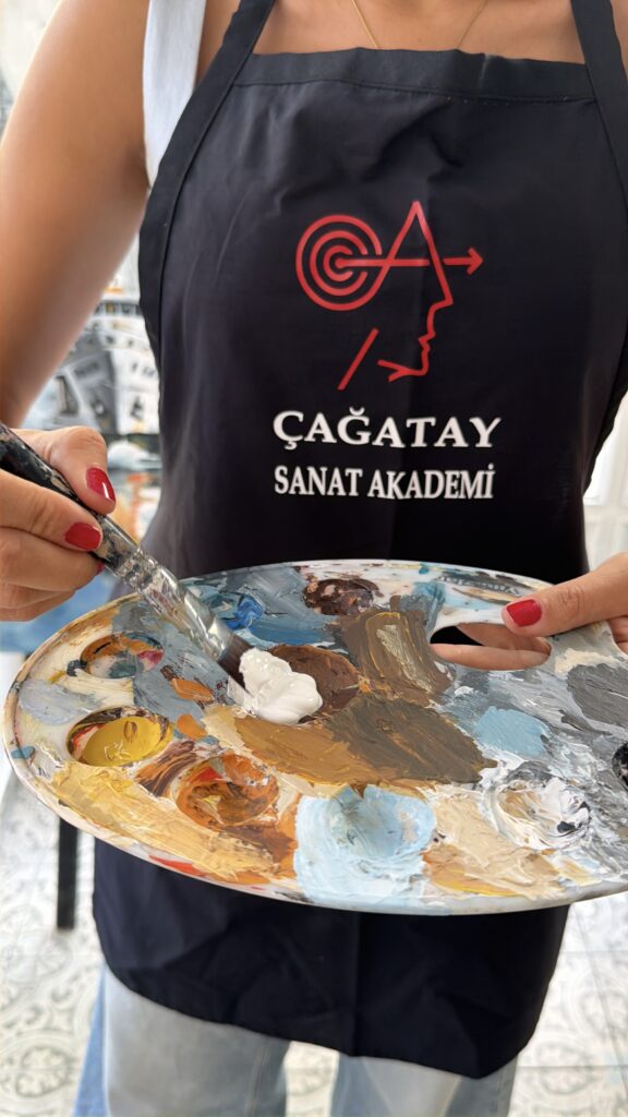 Çağatay Sanat Akademi önlüğü giyen kişinin palet üzerinde fırçayla boya karıştırması